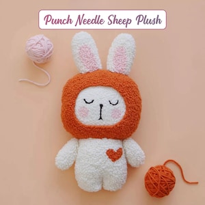 Può includere: Un peluche di pecora bianco e arancione fatto a mano con un cuore sul petto e la scritta "Punch Needle Sheep Plush". La pecora ha una testa rotonda arancione, orecchie bianche con interno rosa e occhi chiusi. Due gomitoli di lana sono accanto al peluche.