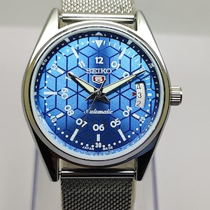 Peut inclure: Une montre-bracelet en acier inoxydable argenté avec un cadran bleu orné d'un motif géométrique en nid d'abeille. Elle possède un bracelet en maille, des aiguilles luminescentes, un guichet de date et l'inscription Seiko 5 Automatic Japan Made.