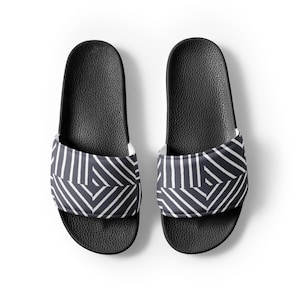 Op de afbeelding: Een paar zwarte slippers met een wit geometrisch patroon op de band. De slippers hebben een getextureerd voetbed en een effen zwarte zool, perfect voor casual gebruik.