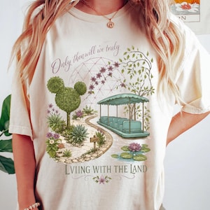 Könnte beinhalten: Cremefarbenes T-Shirt mit einer skurrilen Gartenszene. Es zeigt ein teal-farbenes Boot, eine Mickey Mouse-förmige Topiary und den Text "Only then will we truly Living with the Land". Das Design umfasst einen Teich mit Seerosen und einen gewundenen Pfad.