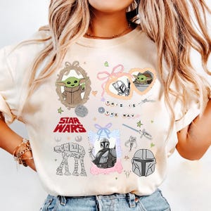 Może przedstawiać: Kremowa koszulka z grafiką Star Wars. Projekty obejmują Grogu, Mandalorianina i tekst "THIS IS THE WAY". Inne grafiki to AT-AT walker, miecz świetlny i Gwiazda Śmierci.
