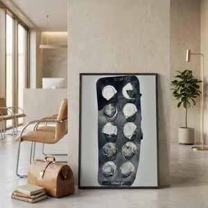 May include: A framed artwork featuring a close-up of a used blister pack of pills. The pills are white, and the packaging is dark gray. The artwork is displayed in a modern interior setting with a leather bag and chairs.