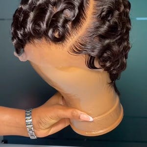 May include: A dark brown wig with finger waves is displayed on a mannequin head. The wig has a side part and is styled with tight, glossy curls. The mannequin head is a light brown color, and a hand with a silver watch is holding the head.