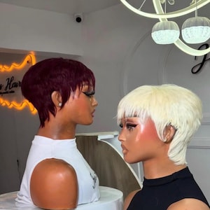 May include: Two mannequin heads with short, stylish haircuts. One has a deep burgundy color, the other is a light blonde. Both mannequins are wearing sleeveless tops. The background includes a neon sign and decorative lighting.