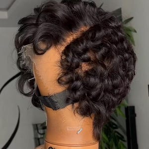 Peut inclure: Perruque courte et bouclée noire, coiffée sur une tête de mannequin. La perruque présente un devant en dentelle et est fixée avec une sangle noire. Le mannequin est de couleur marron clair. Les boucles de la perruque sont serrées et créent un look volumineux.