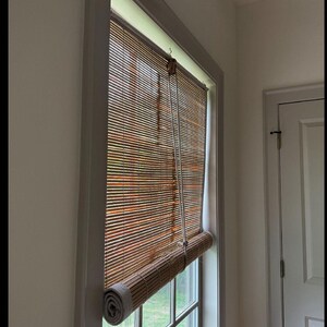 Könnte beinhalten: Ein Bambus-Rollo ist teilweise in einem Fenster heruntergelassen. Das Rollo hat eine warme braune Farbe und ist unten aufgerollt. Der Fensterrahmen ist weiß und die Wand ist hellgrau.