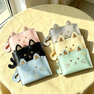 May include: A collection of six rectangular coin purses in pastel and black colours, each featuring cat ears with gold glitter accents, a printed cat face with whiskers, and a dangling cat tail zipper pull. Two purses feature the letters QE and BP.