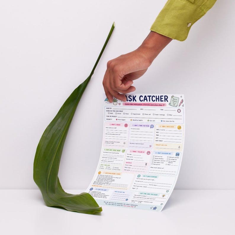 May include: A Task Catcher planning sheet with a white background, colorful text, and various fillable sections for project management, featuring a long, curved green leaf positioned vertically next to the paper document.