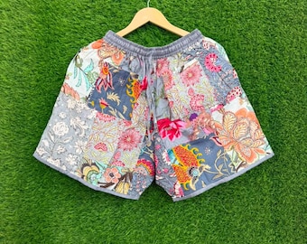 Puede incluir: Unos pantalones cortos con diseño de patchwork que presentan varios estampados florales y botánicos en tonos azul, rosa, naranja y blanco. Los pantalones cortos tienen una cintura elástica con cordón y se muestran en una percha de madera.