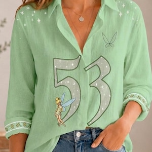 May include: A light green button-down shirt featuring a large number 53 graphic in a glittery silver font, accented with small white stars, a small fairy illustration, and a delicate butterfly outline on the chest, paired with blue denim jeans.