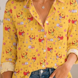 May include: A long-sleeve button-down shirt in a bright yellow hue featuring an all-over print of Winnie the Pooh characters, small pink flowers, and honey pots. The shirt has a classic collar, a front button placket, and cuffed sleeves.