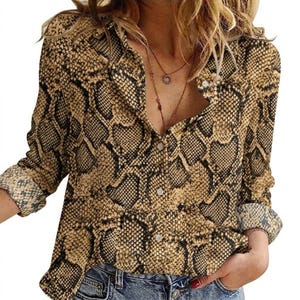 May include: A button-down shirt with a snakeskin pattern in shades of brown and black. The long-sleeved shirt has a collared neckline and buttoned cuffs. The shirt is unbuttoned at the top, revealing a glimpse of a necklace.