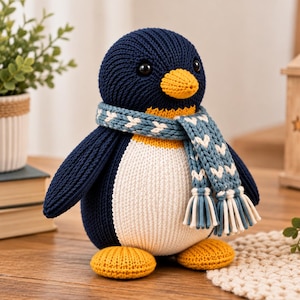 May include: A knitted penguin plush toy featuring a dark navy blue body, a white belly, and bright yellow feet and beak. The penguin wears a light blue scarf with a white chevron pattern and fringed ends. It has two small black plastic eyes.