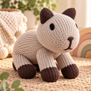 Könnte beinhalten: Ein gestricktes Stofftier in Form einer Katze mit hellbeigem Körper sowie dunkelbraunen Ohren, Pfoten und Schwanz. Die Katze hat schwarze gestickte Augen sowie eine dunkelbraun gestickte Nase und Mund mit einer durchgehenden Rippstruktur.