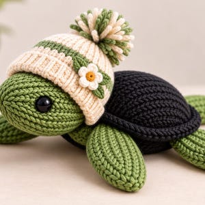 Pode incluir: Um brinquedo de tartaruga em crochê com cabeça e membros verdes, carapaça preta e um gorro de malha creme com listras verdes, um pompom grande no topo e uma pequena aplicação de flor branca com centro amarelo na lateral do gorro.