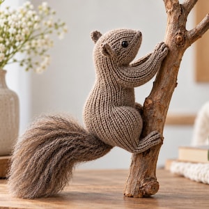 Puede incluir: Una figura de ardilla tejida en color marrón claro con una cola texturizada y esponjosa, ojos pequeños y oscuros y orejas cosidas, posicionada como si estuviera trepando por una rama de madera vertical. El cuerpo muestra puntadas de tejido visibles.