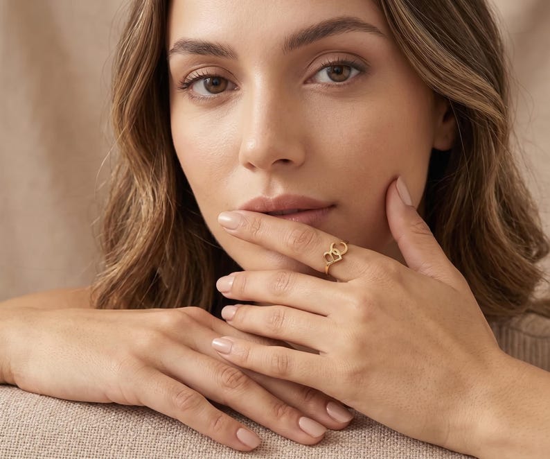 K&ouml;nnte beinhalten: Ein goldener Ring mit zwei ineinandergreifenden, herzf&ouml;rmigen Umrissen. Die Ringschiene ist d&uuml;nn und poliert und zeigt ein schlichtes Design, das am Finger anliegt. Das Metall hat einen warmen, reflektierenden Gelbgoldton, der das Licht einf&auml;ngt.
