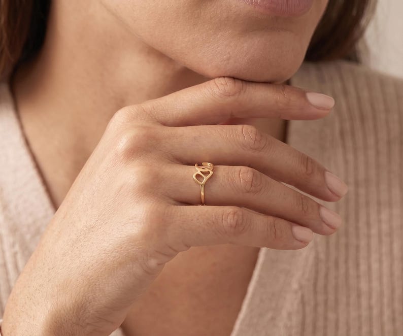 K&ouml;nnte beinhalten: Ein goldener Ring mit einem filigranen, durchbrochenen Design am Ringband. Der Blickfang besteht aus zwei miteinander verbundenen, abgerundeten, organischen Formen, die horizontal auf dem Finger liegen und ein flie&szlig;endes, poliertes Aussehen erzeugen.
