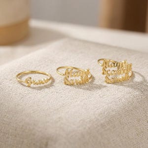 May include: Three gold-toned metal rings featuring personalized script lettering. The first ring displays the name Grace, the second ring features the names May and Sarah, and the third ring showcases the names Maya, Scarlett, and Sarah in a stacked design.