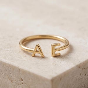 May include: A gold-toned open ring featuring two capital letters, A and E, positioned at the ends of the band. The ring has a smooth, polished finish and a simple, circular design that remains open between the two letters.