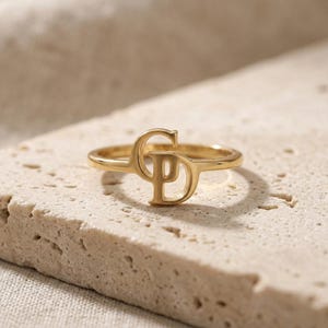 Op de afbeelding: Een goudkleurige ring met een centraal monogramontwerp waarin de letters C en P met elkaar zijn verweven. De ringband is dun en cirkelvormig en heeft een gepolijste metalen afwerking die het licht reflecteert op de letters en de ringband.