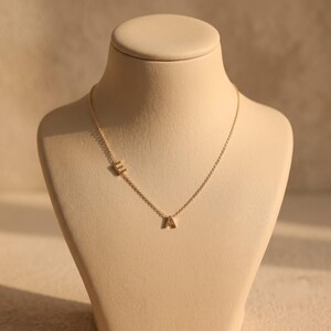 May include: A gold-toned necklace featuring a delicate chain with two small, uppercase letter charms, E and A, positioned asymmetrically along the length of the chain. The letter E is placed higher on the left side, while the letter A sits at the center.