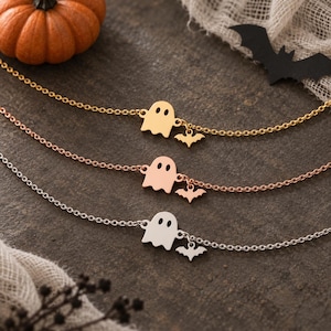 May include: Three delicate chain bracelets in gold, rose gold, and silver tones. Each bracelet features a small ghost-shaped charm and a tiny bat-shaped charm attached to the chain, creating a layered look with a Halloween theme.