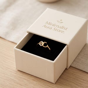 K&ouml;nnte beinhalten: Ein goldfarbener Ring mit den Buchstaben D und C, pr&auml;sentiert in einer cremefarbenen Schmuckschatulle zum Schieben mit schwarzem Samtinnenfutter. Auf dem Deckel steht der Text Minimalist Aura Store sowie ein kleines Sternsymbol &uuml;ber einer Hand.