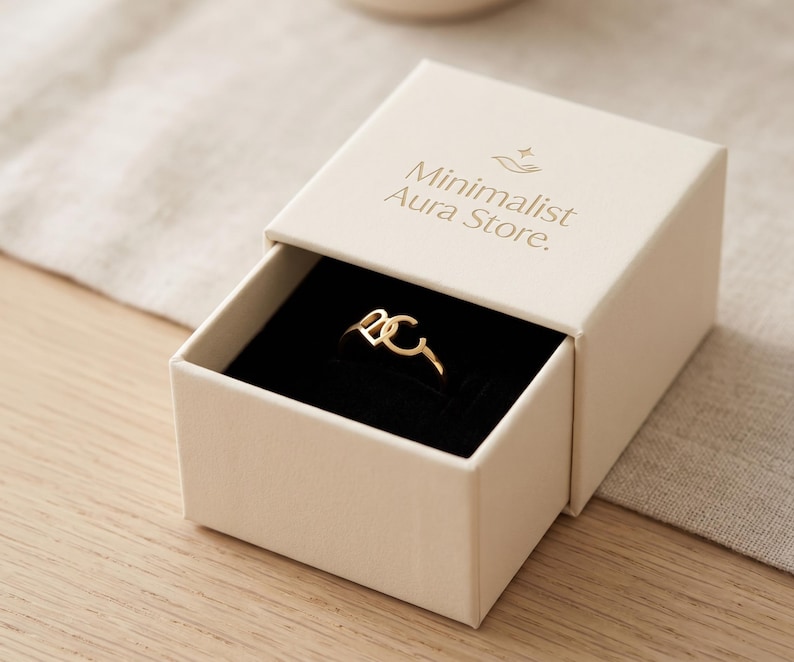 K&ouml;nnte beinhalten: Ein goldfarbener Ring mit den Buchstaben D und C, pr&auml;sentiert in einer cremefarbenen Schmuckschatulle zum Schieben mit schwarzem Samtinnenfutter. Auf dem Deckel steht der Text Minimalist Aura Store sowie ein kleines Sternsymbol &uuml;ber einer Hand.