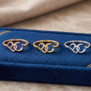 K&ouml;nnte beinhalten: Drei Ringe in einer Reihe angeordnet, jeder mit einem Design aus zwei ineinandergreifenden Herzformen. Die Ringe sind in drei verschiedenen Metallt&ouml;nen gehalten: Ros&eacute;gold, Gelbgold und Silber, pr&auml;sentiert auf einer blauen Samtunterlage.