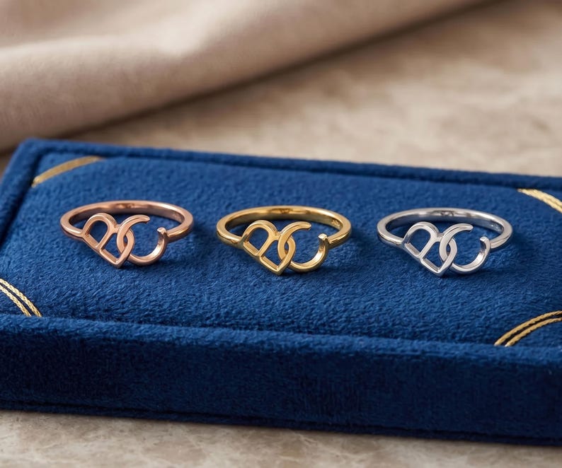 K&ouml;nnte beinhalten: Drei Ringe in einer Reihe angeordnet, jeder mit einem Design aus zwei ineinandergreifenden Herzformen. Die Ringe sind in drei verschiedenen Metallt&ouml;nen gehalten: Ros&eacute;gold, Gelbgold und Silber, pr&auml;sentiert auf einer blauen Samtunterlage.