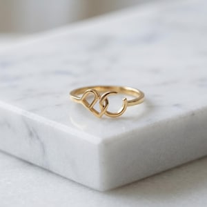 May include: A gold ring featuring two interconnected letters, D and C, forming the focal point of the band. The metal has a polished finish, and the design consists of a capital letter D linked to a capital letter C, creating a continuous loop.