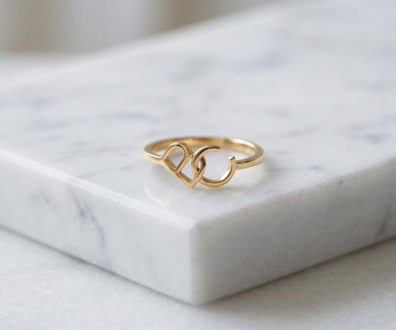 K&ouml;nnte beinhalten: Ein goldener Ring mit zwei miteinander verbundenen Buchstaben, D und C, die den Mittelpunkt des Bandes bilden. Das Metall hat eine polierte Oberfl&auml;che und das Design besteht aus einem Gro&szlig;buchstaben D, der mit einem Gro&szlig;buchstaben C verbunden ist.