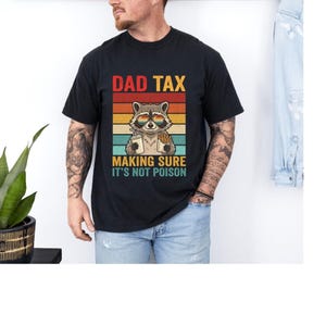A man wearing a black t-shirt with a graphic design of a raccoon wearing sunglasses and holding a sign that says "Making sure it's not poison".