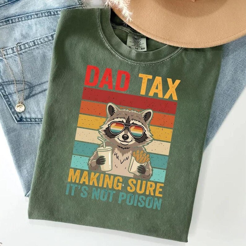 A green t-shirt with a graphic design featuring a raccoon wearing sunglasses and holding a drink, along with the text "DAD TAX MAKING SURE IT'S NOT POISON".