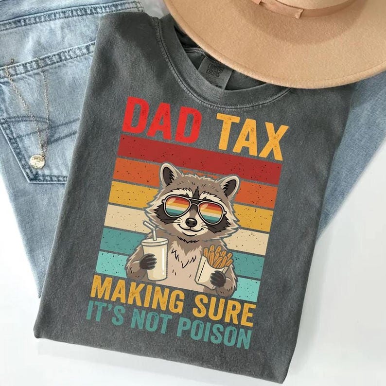 A gray t-shirt with a graphic design featuring a raccoon wearing sunglasses and holding a drink, along with the text "DAD TAX MAKING SURE IT'S NOT POISON".