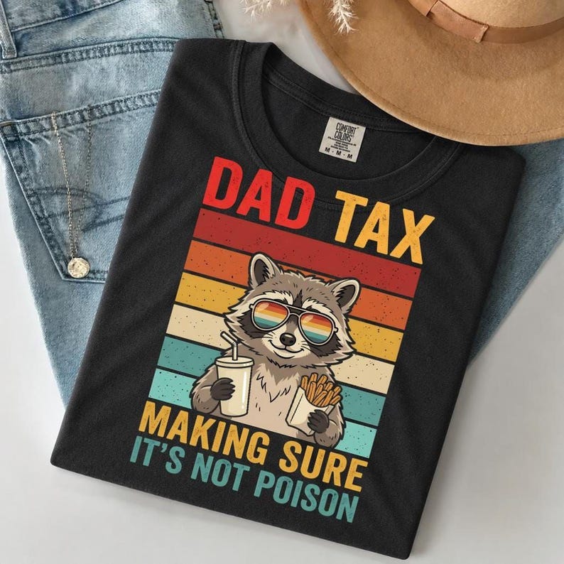 A black t-shirt with a graphic design featuring a raccoon wearing sunglasses and holding a cup of fries, along with the text "DAD TAX MAKING SURE IT'S NOT POISON".