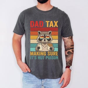 A man wearing a gray t-shirt with a graphic design of a raccoon wearing sunglasses and holding a sign that says "DAD TAX MAKING SURE IT'S NOT POISON".