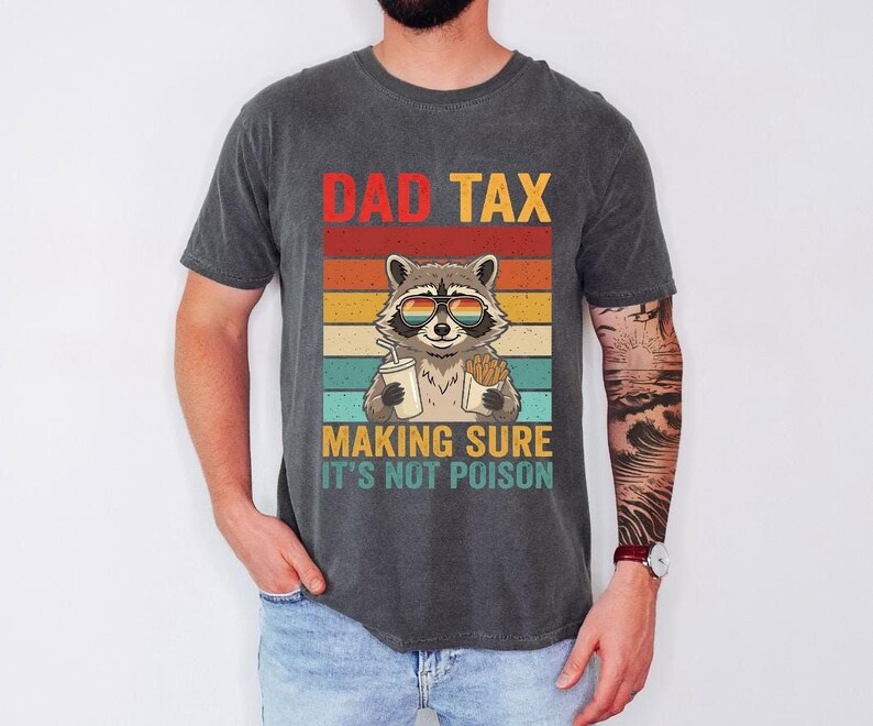 A man wearing a gray t-shirt with a graphic design of a raccoon wearing sunglasses and holding a sign that says "DAD TAX MAKING SURE IT'S NOT POISON".