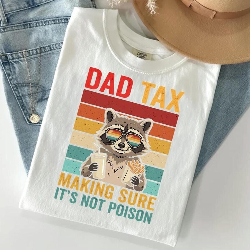 A white t-shirt with a graphic design featuring a raccoon wearing sunglasses and holding a sign that says "DAD TAX MAKING SURE IT'S NOT POISON".