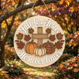 May include: A round wooden hanging sign featuring a central wooden cross, three pumpkins, and scattered autumn leaves. The text reads Fall for Jesus He never leaves in white script. The sign is suspended by a thin rope against a blurred outdoor background.