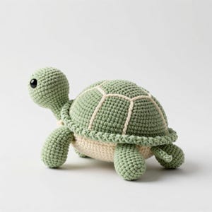 Puede incluir: Un juguete de tortuga tejido a ganchillo con un caparazón verde salvia con detalles cosidos en color crema, una cabeza verde salvia a juego y un ojo de plástico negro liso. La parte inferior de la tortuga es de color crema sólido y las cuatro patas son texturizadas.