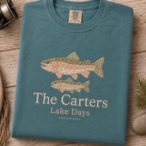 May include: A bay-colored crew neck t-shirt featuring an embroidered design of two trout fish above the text The Carters and Lake Days in a light font. The shirt has a Comfort Colors brand label at the inner neckline indicating a size large.