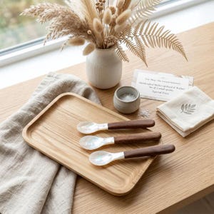 Puede incluir: Una bandeja de madera contiene tres cucharas de nácar con mangos de madera oscura. Un pequeño cuenco de cerámica con sal, una servilleta de lino con un diseño de hojas y un jarrón con flores secas también están sobre la mesa de madera.