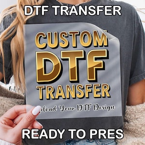May include: A gray DTF transfer sheet with the words "DTF TRANSFER" at the top and bottom. The center of the sheet has the words "CUSTOM DTF TRANSFER" in gold lettering. The text "Upload Your DTF Design" is also visible.