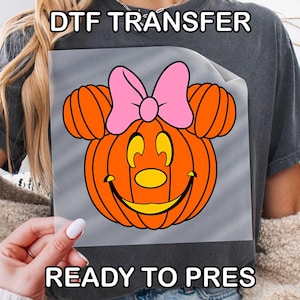 May include: A DTF transfer featuring a cartoon pumpkin design with Mickey Mouse ears and a pink bow. The pumpkin is orange with a yellow face and black outlines. The text "DTF TRANSFER" and "READY TO PRESS" are visible.