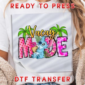 May include: White t-shirt featuring a colorful graphic with the words "Vacay Mode" and a cartoon character. The letters "M" and "E" are pink with a heart pattern. The text "READY TO PRESS" and "DTF TRANSFER" are in red.