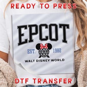 May include: White t-shirt featuring a graphic design with the word "EPCOT" in black block letters. Below the word "EPCOT" is a Minnie Mouse head with a red bow. The text "EST. 1982" and "WALT DISNEY WORLD" are also included.