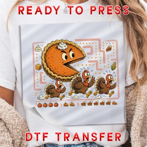 May include: A white t-shirt featuring a DTF transfer graphic of a pumpkin pie character chasing three turkeys in a maze-like design. The pie has a dollop of whipped cream on top, and the turkeys appear startled. Small food icons and orange lines complete the scene.