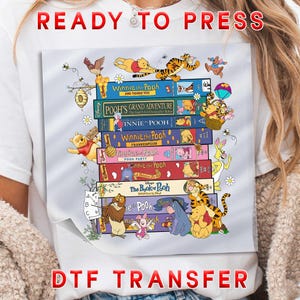 May include: A white t-shirt featuring a colorful graphic of stacked Winnie the Pooh books. The books are adorned with illustrations of Pooh, Tigger, Piglet, and other characters. The words "READY TO PRESS" and "DTF TRANSFER" are printed on the shirt.